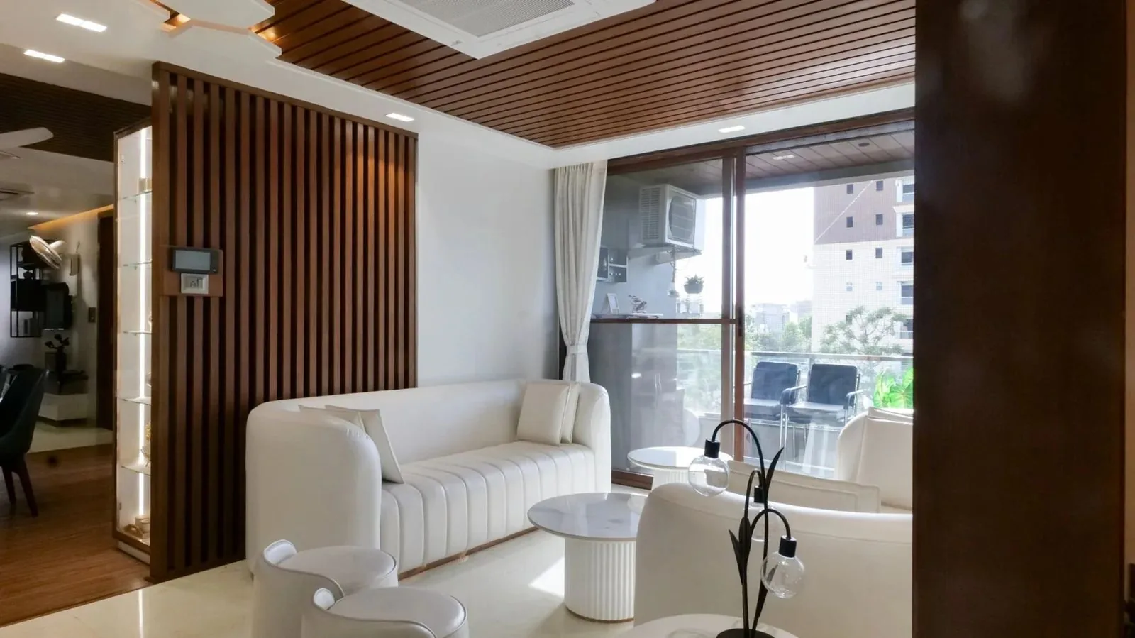 Modern living room with a fluted wood partition, a wooden slatted ceiling, white curved sofas and balcony doors