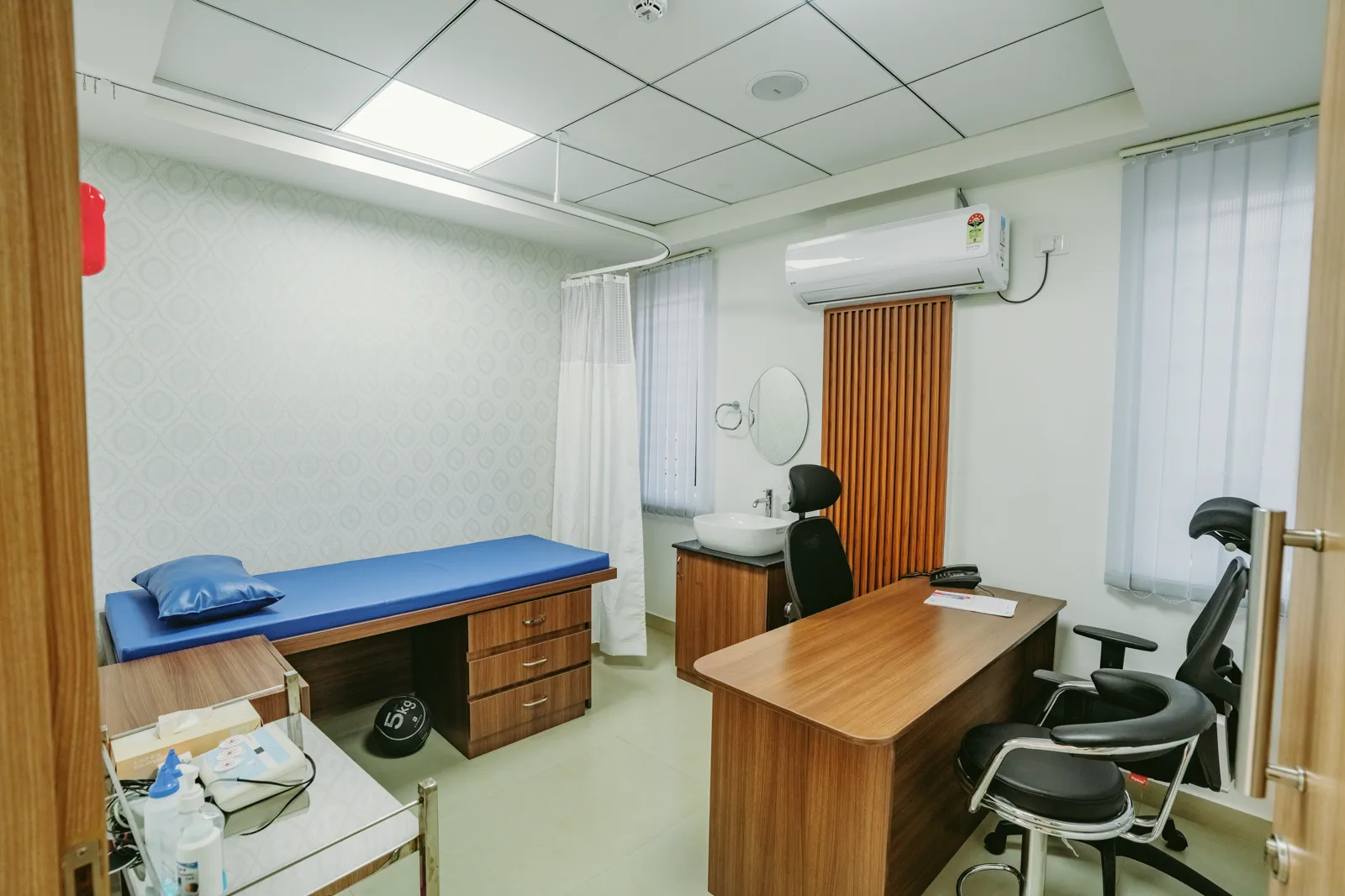 Consultation room by Candidscape at Apollo Clinic, Akshay Nagar, Bangalore, with a wooden doctor's desk, an examination couch and a hand wash basin