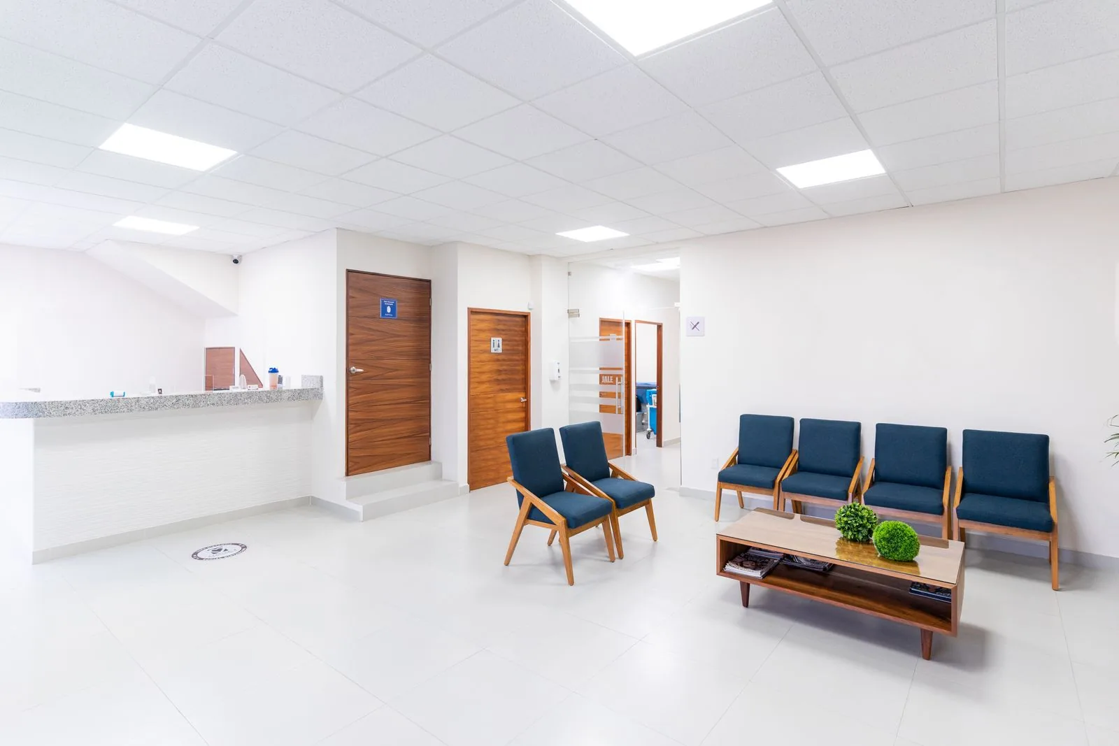 Bright clinic waiting room with blue upholstered chairs, a wooden coffee table, a reception counter and consultation room doors