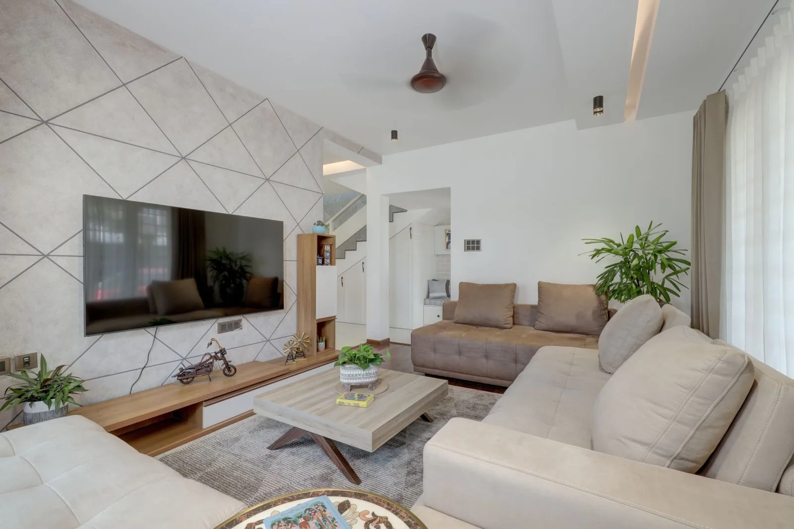 Modern apartment living room with a geometric-panelled TV wall, neutral L-shaped sofas, a stone-top coffee table and indoor plants